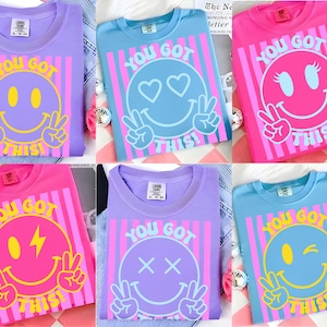 Peut inclure: Plusieurs t-shirts colorés avec un motif de smiley et le texte "YOU GOT THIS!". Les t-shirts sont disponibles en rose, bleu et violet, chacun avec une expression de smiley différente et des accents.