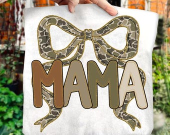 May include: White t-shirt featuring the word "MAMA" in brown and tan bubble letters. A camouflage bow design is above the word "MAMA". The shirt is held up by a hand.