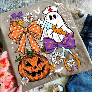 May include: A tan t-shirt featuring a Halloween-themed graphic. The design includes a smiling jack-o'-lantern, a ghost wearing a nurse's cap and holding a syringe, bows, daisies, and bats. The shirt is decorated with orange, purple, and white accents.