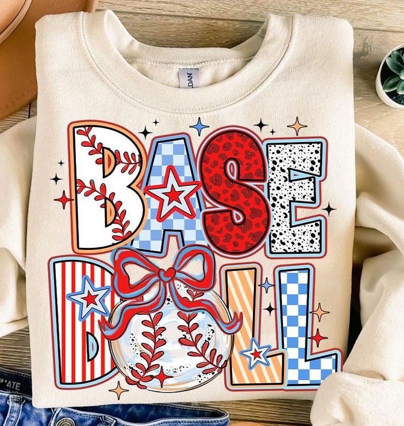May include: A cream-colored sweatshirt with a colorful graphic design that says "BASEBALL" with a baseball and a bow. The graphic has red, white, blue, and black colors.