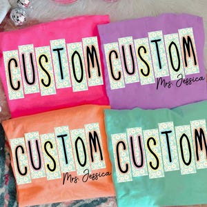 Puede incluir: Cuatro camisetas coloridas en rosa, morado, naranja y turquesa. Cada camiseta presenta la palabra "CUSTOM" en negrita, en negro, con un estampado de margaritas. El nombre "Mrs. Jessica" está impreso en dos de las camisetas.