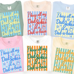 May include: Six t-shirts in different colours, each displaying the text "I keep all my Dad Jokes in a Dad-a-base." The design incorporates stars and a graphic of a data storage device. Colours include green, pink, white, blue, and yellow.