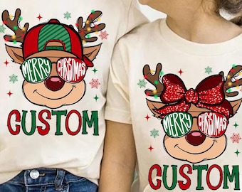May include: Two cream-coloured t-shirts featuring a Christmas reindeer design. The reindeer wears sunglasses with "Merry Christmas" text, a red and green hat, and antlers with lights. The word "CUSTOM" is printed below the reindeer.