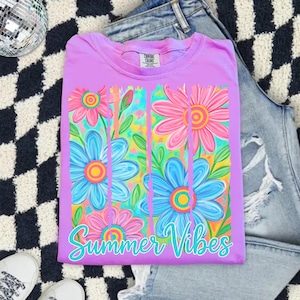 May include: A lilac-colored t-shirt with a floral design featuring pink and blue flowers. The shirt has the words "Summer Vibes" in a turquoise script. The shirt is folded on a black and white checkered surface.