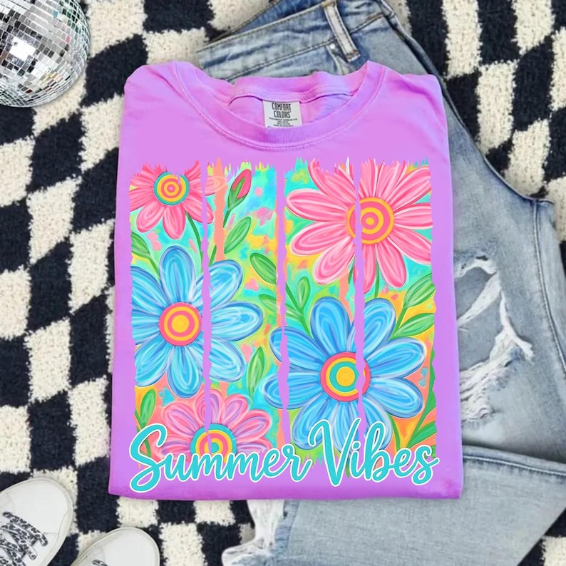 May include: A lilac-colored t-shirt with a floral design featuring pink and blue flowers. The shirt has the words "Summer Vibes" in a turquoise script. The shirt is folded on a black and white checkered surface.
