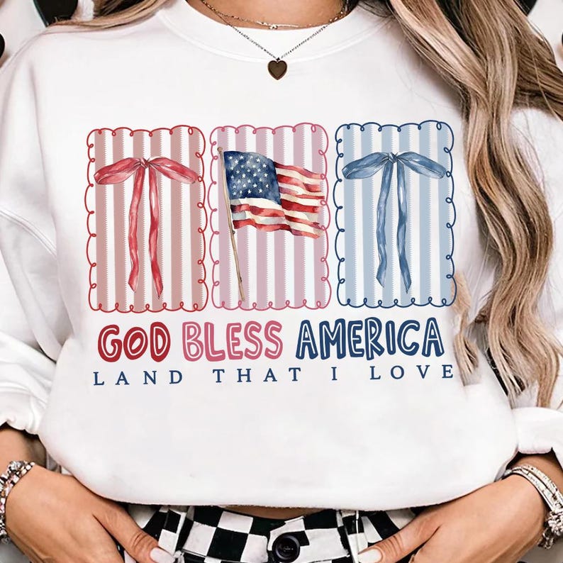 May include: White sweatshirt with a patriotic design. Features three rectangular frames with bows and an American flag. The text "GOD BLESS AMERICA" and "LAND THAT I LOVE" are printed below the frames.