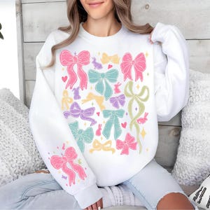 May include: White sweatshirt with a colorful bow pattern. The bows are in shades of pink, purple, green, and yellow. There are also small bunny silhouettes scattered throughout the design.