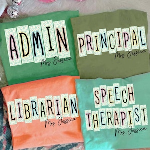 May include: Four brightly coloured t-shirts, each displaying a different job title: "ADMIN", "PRINCIPAL", "LIBRARIAN", and "SPEECH THERAPIST". The shirts are in green, olive green, coral, and teal. Each shirt has the name "Mrs. Jessica" below the title.