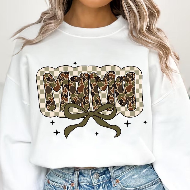 May include: White sweatshirt featuring the word "MAMA" in a camouflage pattern, framed by a tan and white checkered border. A green bow sits beneath the word, with black star accents.