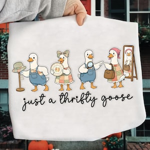 Puede incluir: Bolso tote blanco con una ilustración de dibujos animados de cuatro gansos vestidos con atuendos vintage, cada uno con un accesorio. El texto "just a thrifty goose" está escrito en cursiva debajo de los gansos.