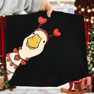May include: Black fabric featuring a whimsical illustration of a goose with a yellow beak and a red heart-shaped headband. The goose is adorned with a garland of red hearts. The design is likely for a t-shirt or similar apparel.