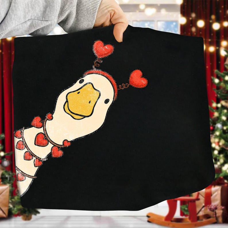 May include: Black fabric featuring a whimsical illustration of a goose with a yellow beak and a red heart-shaped headband. The goose is adorned with a garland of red hearts. The design is likely for a t-shirt or similar apparel.