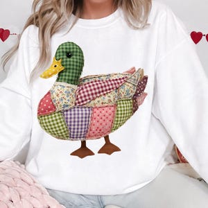 May include: White sweatshirt featuring a patchwork duck design. The duck has a green checkered head, a yellow beak, and a body composed of various patterned fabrics in pink, purple, and floral prints. Brown feet complete the whimsical design.