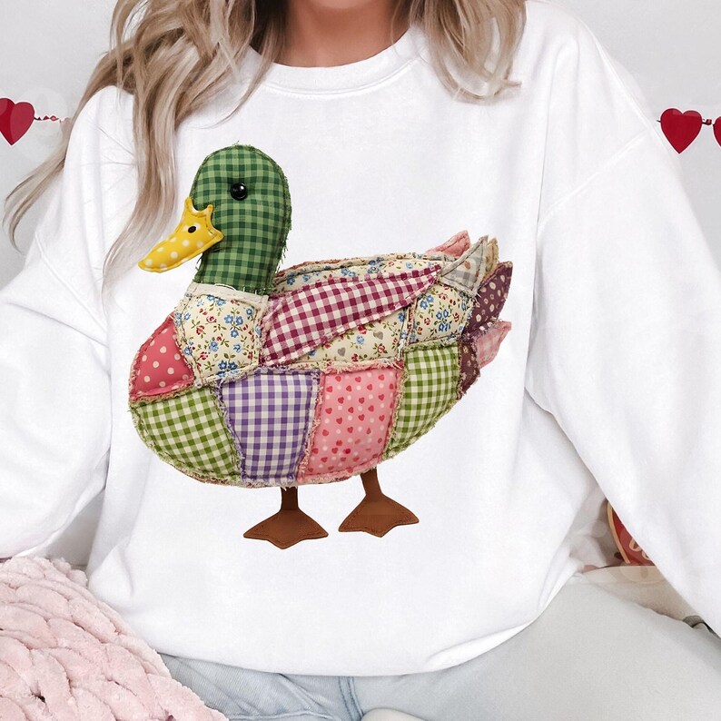 May include: White sweatshirt featuring a patchwork duck design. The duck has a green checkered head, a yellow beak, and a body composed of various patterned fabrics in pink, purple, and floral prints. Brown feet complete the whimsical design.