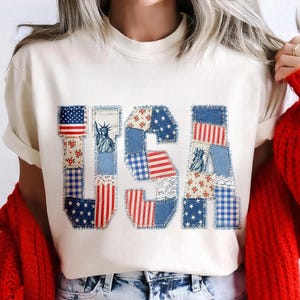 K&ouml;nnte beinhalten: Cremefarbenes T-Shirt mit dem Wort "USA" in einem Patchwork-Design. Die Buchstaben zeigen Muster der amerikanischen Flagge, Sterne und die Freiheitsstatue. Das Shirt hat kurze &Auml;rmel und einen Rundhalsausschnitt.