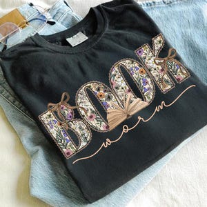 K&ouml;nnte beinhalten: Schwarzes T-Shirt mit dem Schriftzug "BOOK worm" in einem floralen Design. Die Buchstaben sind mit bunten Blumen und Bl&auml;ttern gef&uuml;llt, mit einer kleinen Grafik eines offenen Buches. Eine hellbraune Schleife ist an den Buchstaben befestigt. Das Shirt liegt auf einer blauen Jeans.