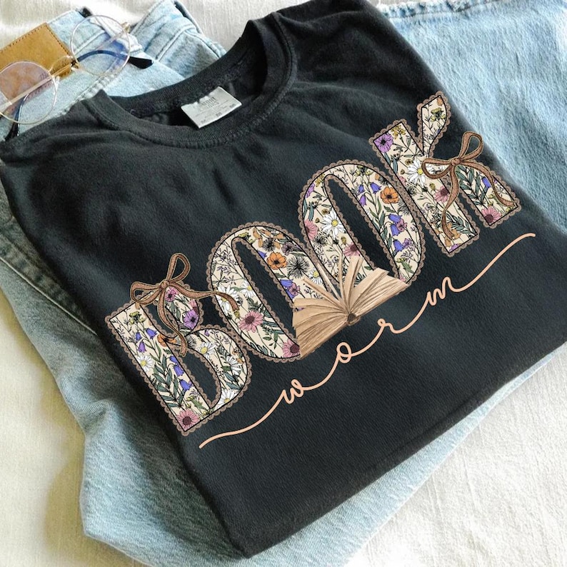 K&ouml;nnte beinhalten: Schwarzes T-Shirt mit dem Schriftzug "BOOK worm" in einem floralen Design. Die Buchstaben sind mit bunten Blumen und Bl&auml;ttern gef&uuml;llt, mit einer kleinen Grafik eines offenen Buches. Eine hellbraune Schleife ist an den Buchstaben befestigt. Das Shirt liegt auf einer blauen Jeans.