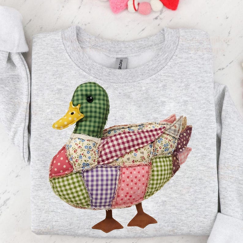 May include: A light gray sweatshirt featuring a patchwork duck design. The duck has a green checkered head, a yellow beak, and a body composed of various fabric patches in pink, purple, and floral patterns. Brown feet complete the whimsical design.