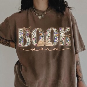 K&ouml;nnte beinhalten: Braunes T-Shirt mit dem Schriftzug "BOOK worm" in floralem Design. Die Buchstaben sind mit Blumen und Bl&auml;ttern gef&uuml;llt, mit einem kleinen offenen Buch in der Mitte. Das Wort "worm" ist kursiv geschrieben.
