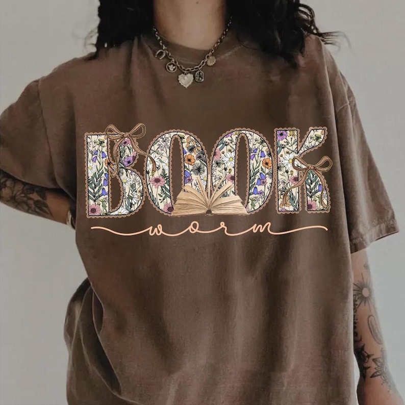 K&ouml;nnte beinhalten: Braunes T-Shirt mit dem Schriftzug "BOOK worm" in floralem Design. Die Buchstaben sind mit Blumen und Bl&auml;ttern gef&uuml;llt, mit einem kleinen offenen Buch in der Mitte. Das Wort "worm" ist kursiv geschrieben.
