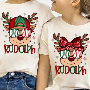 May include: Two off-white t-shirts with a Christmas-themed graphic. The graphic features a cartoon reindeer wearing sunglasses that say "RUN RUN" and the name "Rudolph" in red and green. The reindeer has antlers and is surrounded by snowflakes.