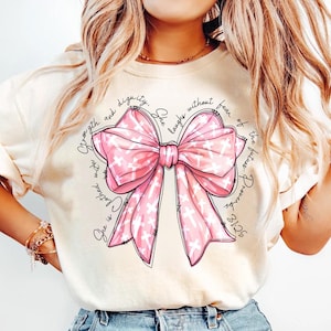 Op de afbeelding: Een wit T-shirt met een roze strik en de tekst "Zij is bekleed met kracht en waardigheid. Zij lacht zonder angst voor de toekomst. Spreuken 31:25"