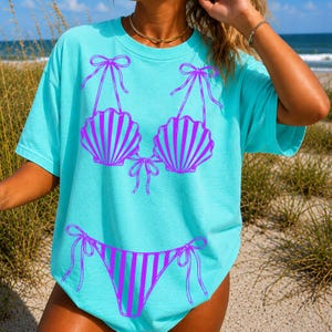 May include: A turquoise t-shirt with a purple seashell bikini design. The bikini top features two seashell cups with bow ties, and the bottom is a striped bikini with bow ties. This is a casual, beach-themed apparel item.