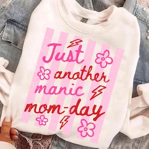 Puede incluir: Sudadera blanca con un gráfico rosa y rojo que dice "Just another manic mom-day". El diseño incluye rayas rosas, flores y rayos. La sudadera se muestra sobre una chaqueta vaquera.