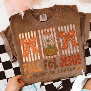 May include: Brown t-shirt featuring a fall design. The graphic includes pumpkins, bows, and the text "FALL FOR JESUS HE NEVER LEAVES" in orange and brown. The shirt has short sleeves and a crew neck.