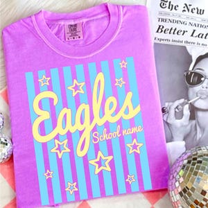 Puede incluir: Una camiseta de color lila con un dise&ntilde;o gr&aacute;fico. El dise&ntilde;o incluye la palabra "Eagles" en escritura amarilla, rayas verticales azules y estrellas amarillas. Las palabras "School name" est&aacute;n debajo.