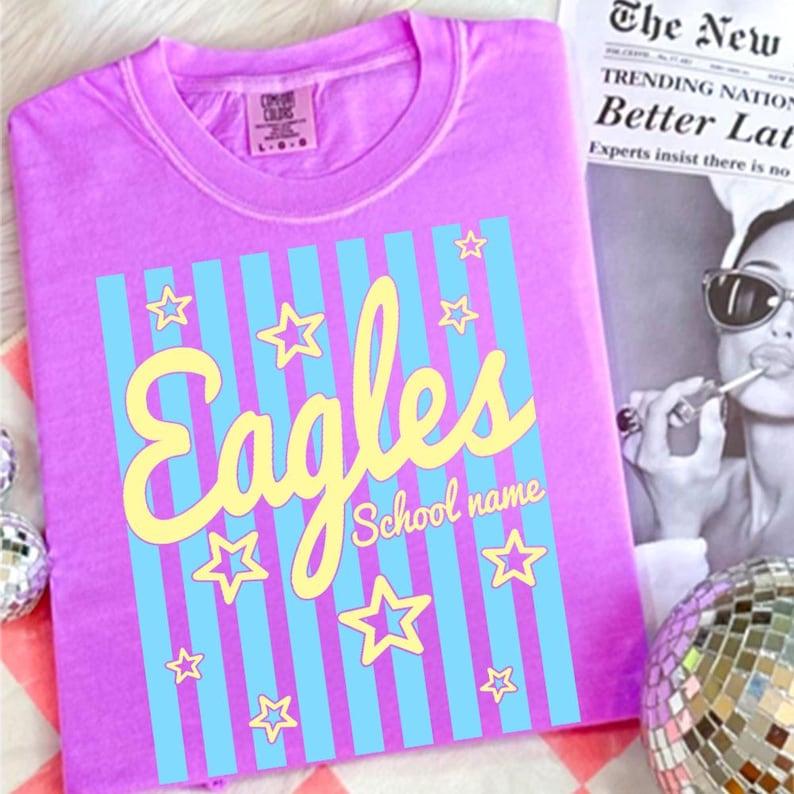 Puede incluir: Una camiseta de color lila con un dise&ntilde;o gr&aacute;fico. El dise&ntilde;o incluye la palabra "Eagles" en escritura amarilla, rayas verticales azules y estrellas amarillas. Las palabras "School name" est&aacute;n debajo.