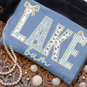 May include: A light blue decorative pillow with the words "LAKE bum" in a pearl and patterned design. The letters are outlined with pearls and filled with various blue and white patterns. A pearl necklace and seashells are in the background.