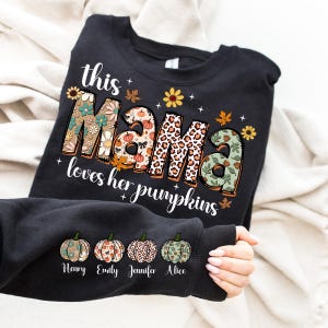 Puede incluir: Sudadera negra con el texto "This Mama loves her pumpkins" en letras de colores con patrones. Cuatro peque&ntilde;as calabazas con los nombres "Henry", "Emily", "Jennifer" y "Alice" est&aacute;n impresas debajo del texto.