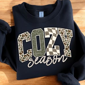 K&ouml;nnte beinhalten: Marineblaues Sweatshirt mit dem Schriftzug "COZY season" in dekorativer Schrift. Die Buchstaben "COZY" haben ein Tarn- und Schachbrettmuster. Das Wort "season" ist in einer Schreibschrift geschrieben.