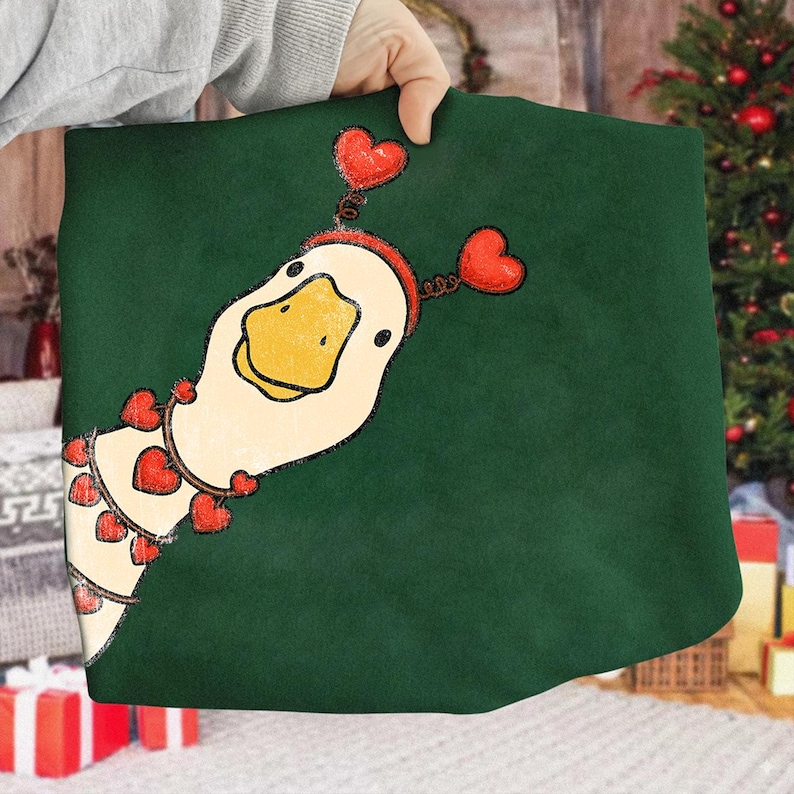 May include: A green fabric item featuring a whimsical goose illustration. The goose has a white head, yellow beak, and is adorned with red heart-shaped accents. The item is held by a person.