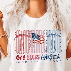 May include: White t-shirt with a patriotic design. The design includes red, white, and blue elements: a waving American flag and decorative bows. The text "GOD BLESS AMERICA LAND THAT I LOVE" is printed below the graphics.