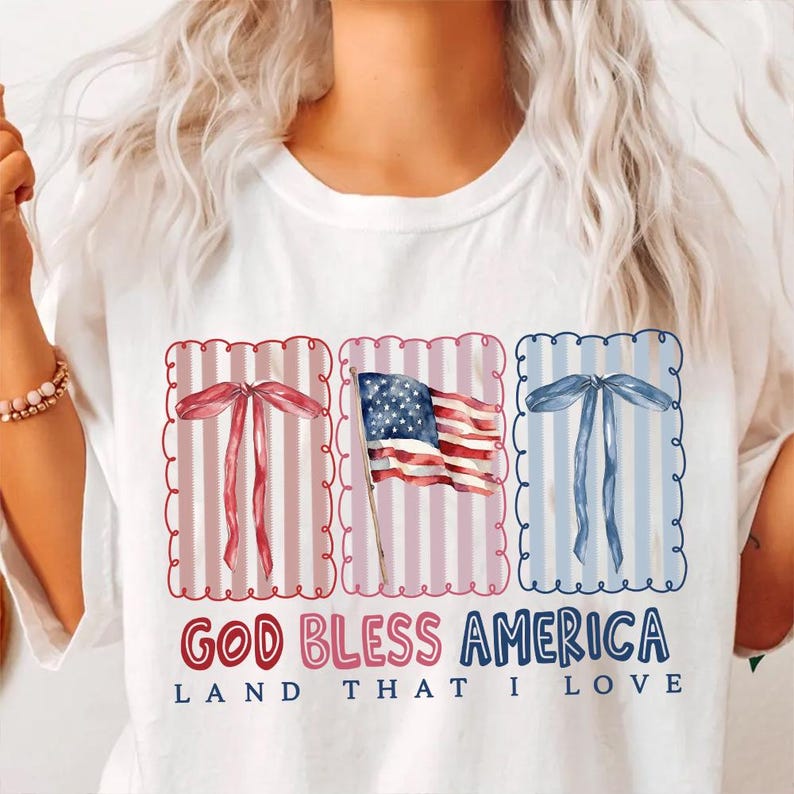 May include: White t-shirt with a patriotic design. The design includes red, white, and blue elements: a waving American flag and decorative bows. The text "GOD BLESS AMERICA LAND THAT I LOVE" is printed below the graphics.