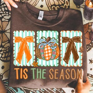 May include: A dark brown sweatshirt featuring three rectangular panels with light teal vertical stripes. The panels display an orange bow, a patterned pumpkin with a light blue bow, and a dark brown bow. The text TIS THE SEASON is printed below.