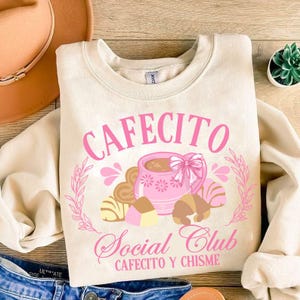 May include: A beige sweatshirt with a pink graphic design that says "Cafecito Social Club Cafecito y Chisme". The graphic features a pink coffee cup with brown and white pastries.