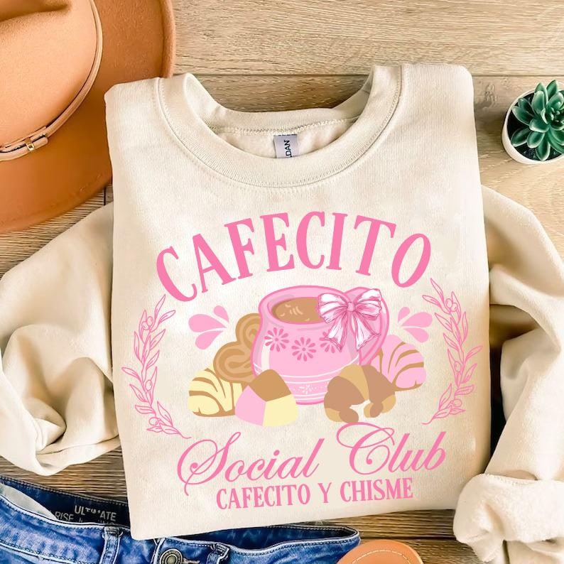 May include: A beige sweatshirt with a pink graphic design that says "Cafecito Social Club Cafecito y Chisme". The graphic features a pink coffee cup with brown and white pastries.