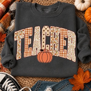 May include: A dark grey crewneck sweatshirt featuring the word TEACHER in large, block-style letters. The letters are filled with a mix of orange and brown floral, plaid, and polka dot patterns. An orange pumpkin with white polka dots sits below.