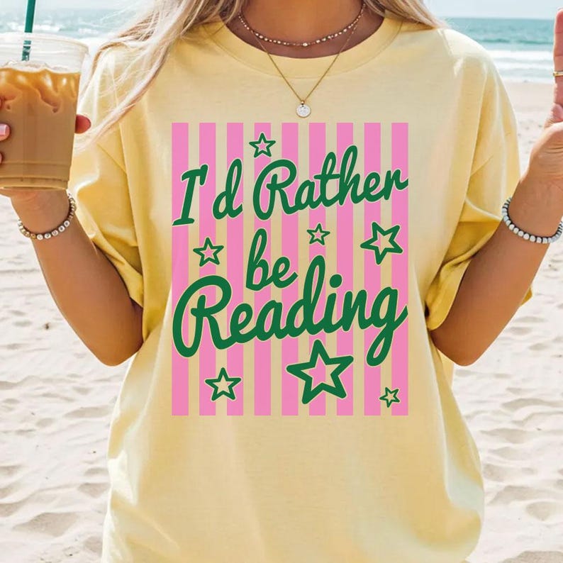 May include: Yellow t-shirt with the text "I'd Rather be Reading" in green script and a pink and white striped background. The shirt has short sleeves and a relaxed fit.