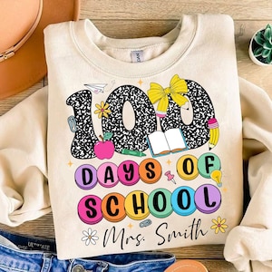 May include: A beige sweatshirt featuring a graphic design of the number 100 made from black and white notebook paper. The number is surrounded by colourful circles that say "100 Days of School" and a bow, book, pencil, and lightbulb. The text "Mrs. Smith" is at the bottom of the design.