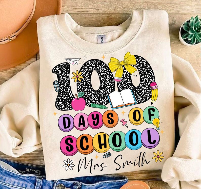 May include: A beige sweatshirt featuring a graphic design of the number 100 made from black and white notebook paper. The number is surrounded by colourful circles that say "100 Days of School" and a bow, book, pencil, and lightbulb. The text "Mrs. Smith" is at the bottom of the design.