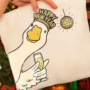 May include: A cream-colored pillow featuring a cartoon goose wearing a gold crown with the words "Happy New Year." The goose is holding a champagne glass, and a disco ball hangs in the background. The design is in black and gold.