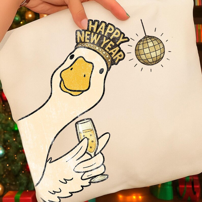 May include: A cream-colored pillow featuring a cartoon goose wearing a gold crown with the words "Happy New Year." The goose is holding a champagne glass, and a disco ball hangs in the background. The design is in black and gold.