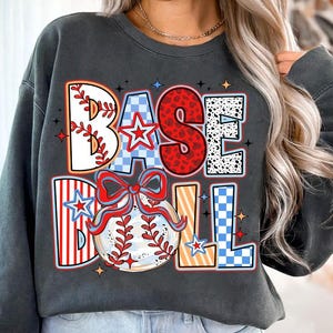 May include: A gray sweatshirt with a colorful graphic design that says "BASEBALL" with a baseball and a bow. The graphic is made up of different patterns like checkered, polka dots, and stripes.