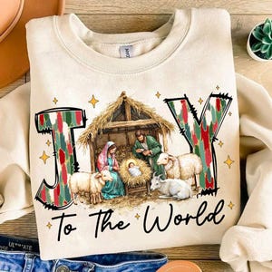 May include: A cream-colored sweatshirt with the word "JOY" in red, green, and white patterned letters. The letters are surrounded by gold stars. The words "To the World" are printed below the letters. A nativity scene with a manger, baby Jesus, Mary, Joseph, and sheep is depicted inside the "O" of the word "JOY".