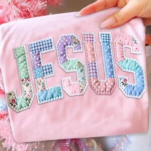 May include: A light pink t-shirt with the word "JESUS" appliqued across the front. The letters are made of patchwork fabric in various colors and patterns, including floral, polka dots, and gingham. The edges of the letters are outlined with white stitching.
