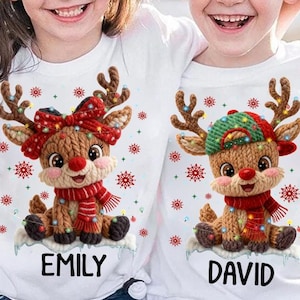 May include: Two white t-shirts with Christmas reindeer designs. One reindeer has a red bow and the name "EMILY" below. The other reindeer has a green and red hat and the name "DAVID" below. Both shirts feature snowflake accents.
